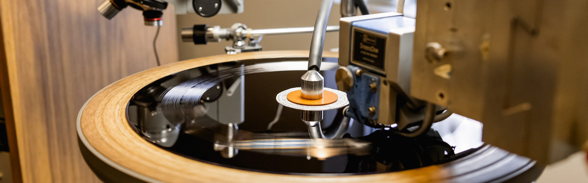 A close-up of a vinyl record being cut by a precision lathe machine, with a focus on the stylus and grooves on the shiny black surface, set against a wood-accented studio background